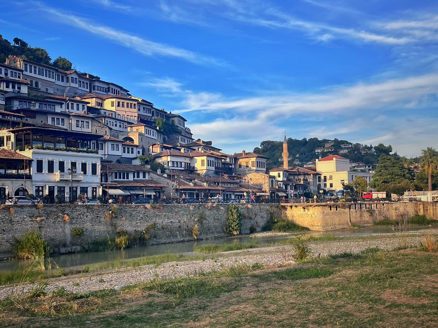 The Albanian town of Berat, with buildings with many windows stacked along a hillside. There is a river in the foreground, and the minaret of a mosque reaches up to the sky